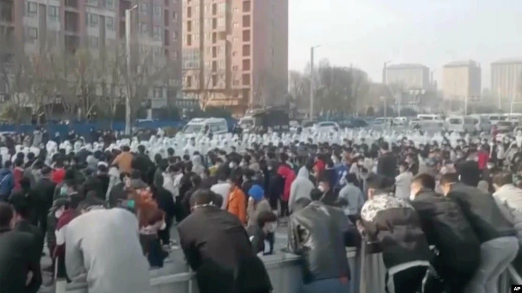 Pengunjuk rasa berhadapan dengan petugas keamanan berpakaian APD putih di kompleks pabrik yang dioperasikan oleh Foxconn Technology Group di Zhengzhou, provinsi Henan, China, 23 November 2022. (AP)