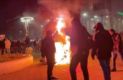 Seorang pria berdiri memegang suar saat para pengunjuk rasa tampak mengamati sebuah motor yang terbakar dalam aksi protes di Rotterdam, Belanda, pada 20 November 2021. (Foto: Video in Verzet via Reuters/Sven Simcic)